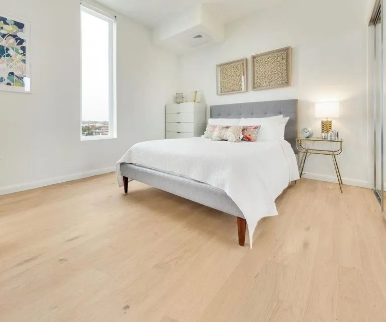 Modern bedroom featuring Royal 14/2mm Oak flooring in a light, natural finish, complemented by a gray upholstered bed and decorative pillows, bright window, and stylish bedside table.
