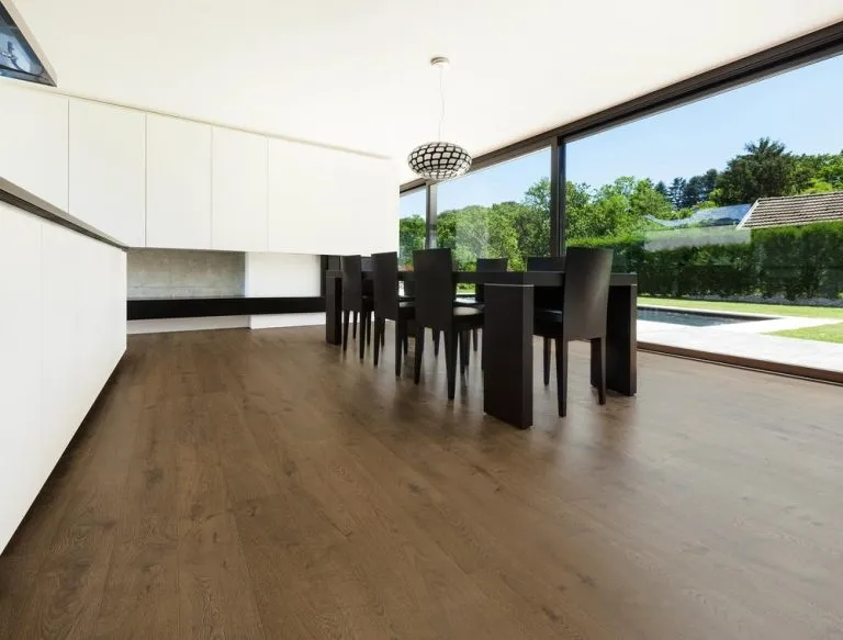 Modern dining area featuring Royal 14/2mm Oak flooring in Mocha, with sleek white cabinetry and large windows overlooking greenery.