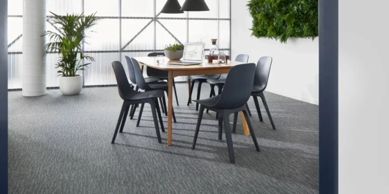 Modern dining area featuring a wooden table surrounded by black chairs, set on textured commercial carpet, with greenery and large windows in the background.