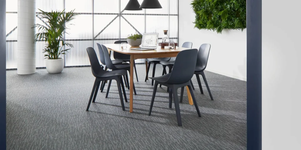 Commercial carpet in a modern office setting with a wooden table, black chairs, and greenery, showcasing durability and style for professional spaces.