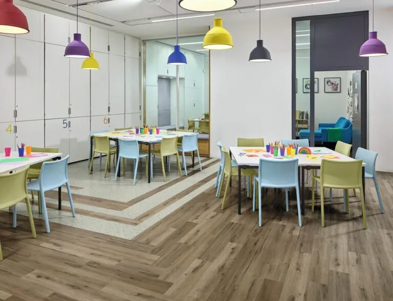 Brightly colored classroom with tables and chairs, featuring Potter Oak luxury vinyl flooring, art supplies on tables, and vibrant pendant lights.